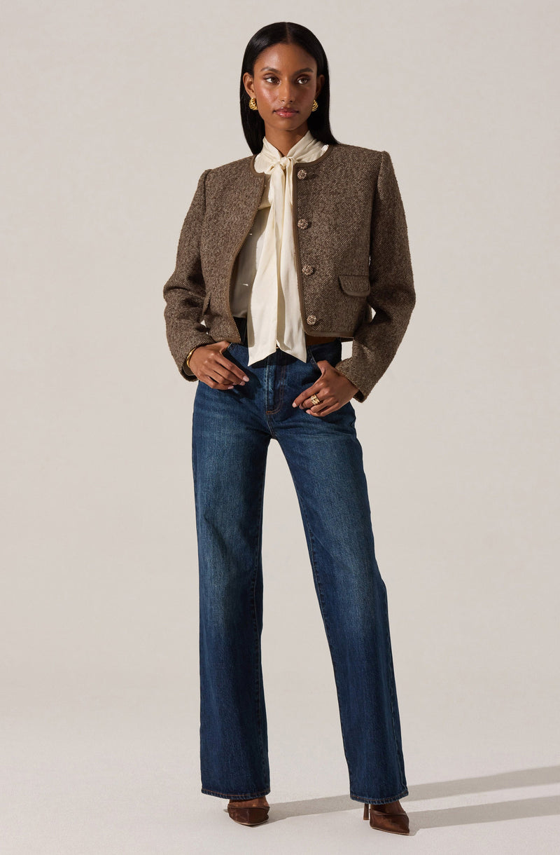 A woman is wearing a brown, textured, cropped blazer with a structured fit and large buttons down the front. Underneath, she has a cream-colored blouse with a bow tied at the collar. She pairs the top with high-waisted, straight-leg blue jeans and brown heeled shoes. She accessorizes with gold earrings and rings.