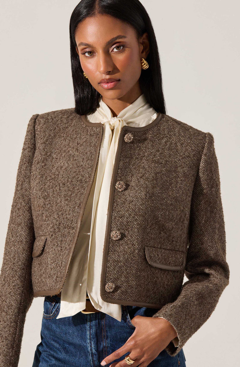 A woman is wearing a cropped, textured brown blazer with a subtle herringbone pattern, featuring a round neckline and four decorative buttons down the front. She pairs it with a cream-colored blouse that has a bow-tie detail at the collar and blue jeans. She accessorizes with gold hoop earrings and rings.