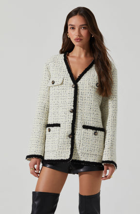 A young woman is wearing a cream-colored, textured woven jacket with black trim along the edges, pockets, and cuffs. The jacket features a single breast pocket with a button and two lower pockets with black piping. She is also wearing black leather shorts and thigh-high black boots.
