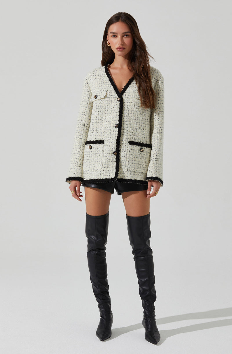 A woman is wearing a white, textured, knit cardigan with black trim along the collar, front opening, cuffs, and pocket edges. The cardigan features large buttons and two flap pockets near the waist. She pairs it with black shorts and thigh-high black leather boots.