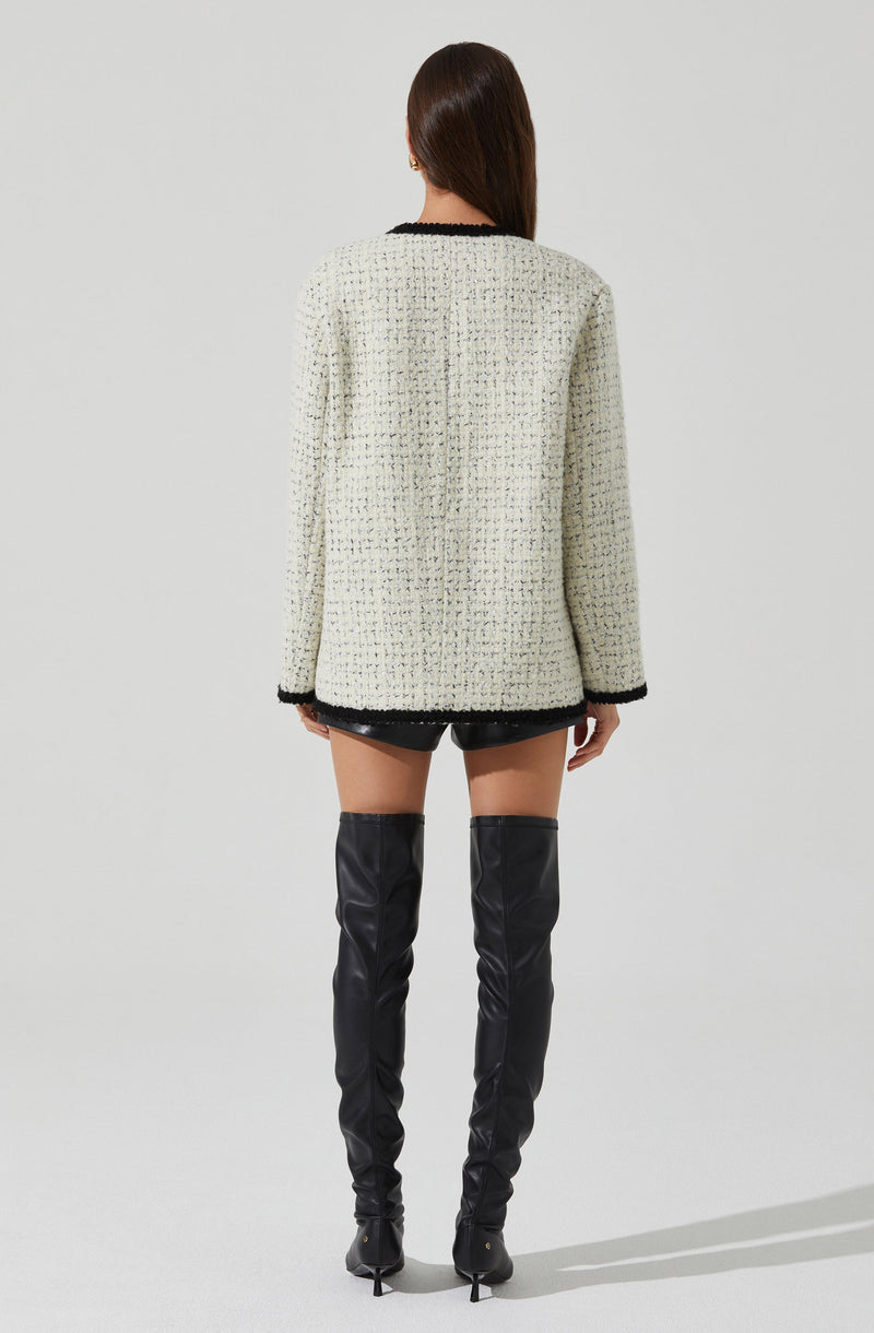 A woman is wearing a cream-colored, textured boucle jacket with black trim around the collar, cuffs, and hem. She is also wearing black shorts and over-the-knee black leather boots with high heels.