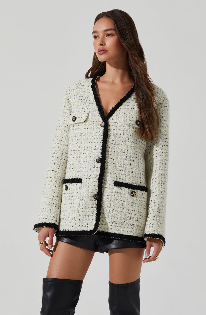 A woman is wearing a cream-colored, textured, tweed-style blazer with black trim along the edges, pockets, and collar. The blazer has two decorative buttons on each pocket and a button-up front. She is also wearing black leather shorts and thigh-high black boots.