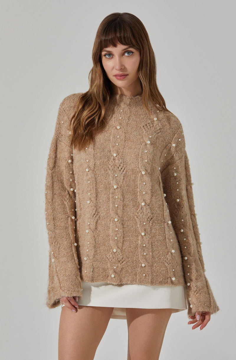 A woman is wearing a loose-fitting, beige cable-knit sweater embellished with vertical rows of small, white pearls. The sweater has a high neckline, long sleeves, and a relaxed fit that extends past her hips. She pairs it with a short, white skirt that is barely visible below the hem of the sweater.