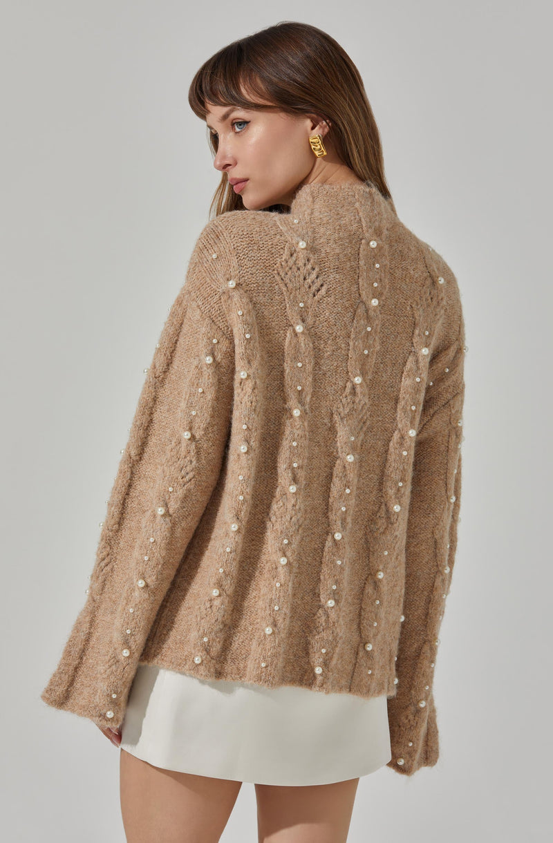 A woman is wearing a beige, fuzzy knit sweater with a high neckline. The sweater features vertical rows of small white pearls or beads embellishing the fabric. She pairs it with a white skirt and accessorizes with large gold earrings.