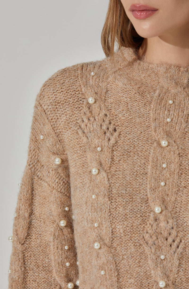 A close-up of a woman wearing a beige knitted sweater embellished with small, round pearl accents scattered across the fabric. The sweater has a textured pattern and a high neckline.