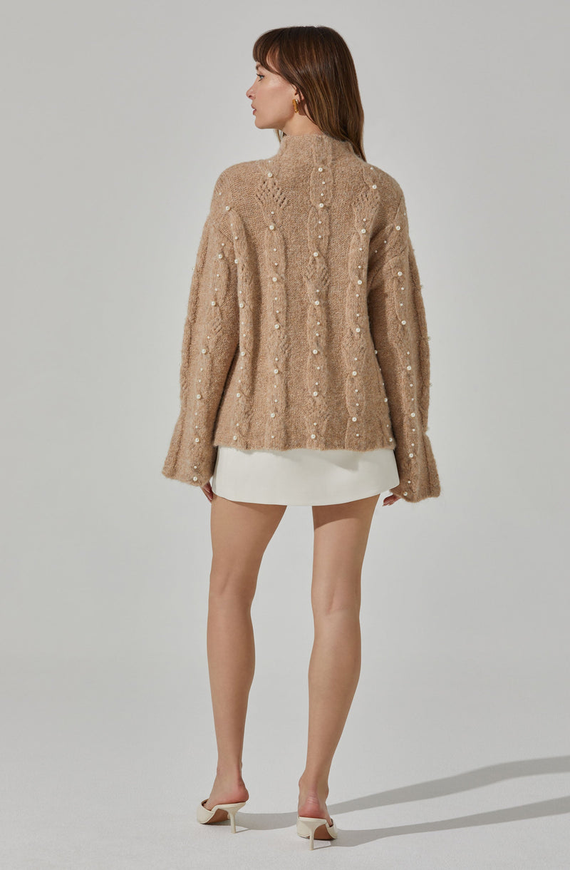 A woman is wearing a beige, chunky knit sweater with a high neckline. The sweater features a textured cable pattern and is decorated with small pearls scattered throughout. She pairs it with a short, white skirt and white high-heeled shoes.