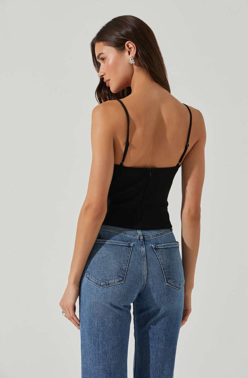 A woman is wearing a black spaghetti strap top that fits snugly and has a back zipper, paired with high-waisted blue jeans with classic five-pocket styling and visible stitching. She has long, dark hair and is accessorizing with large earrings.