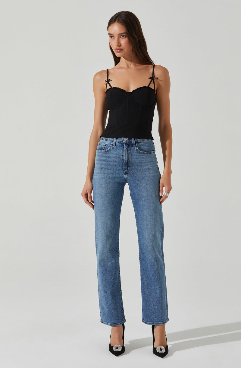 A woman is wearing a black fitted top with thin spaghetti straps, featuring decorative bows on the straps and a structured, corset-like design with sewn details. She pairs this top with high-waisted, straight-leg blue jeans and black high heels with a decorative embellishment on the toe.