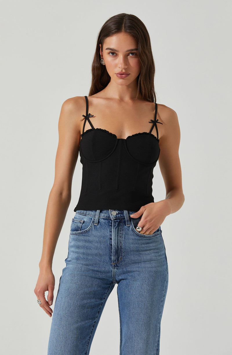 A woman is wearing a fitted black sleeveless top with thin spaghetti straps that feature small bow details at the shoulders. The top has a structured, busty design with visible seams and a slightly scalloped edge along the neckline. She pairs it with high-waisted, light blue jeans.