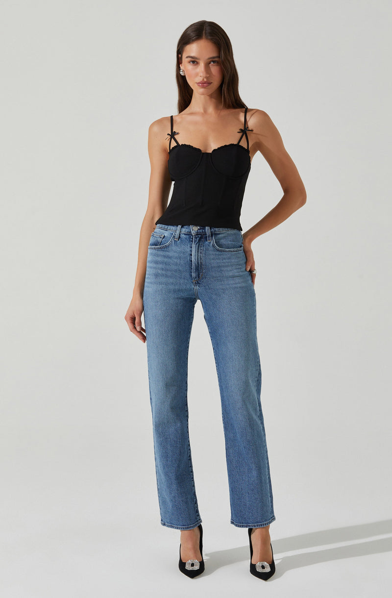 A woman is wearing a black sleeveless top with thin spaghetti straps that have small bows tied at the shoulders. The top has a fitted underwire design with a scalloped neckline. She is also wearing high-waisted, straight-leg blue jeans with a classic five-pocket style.