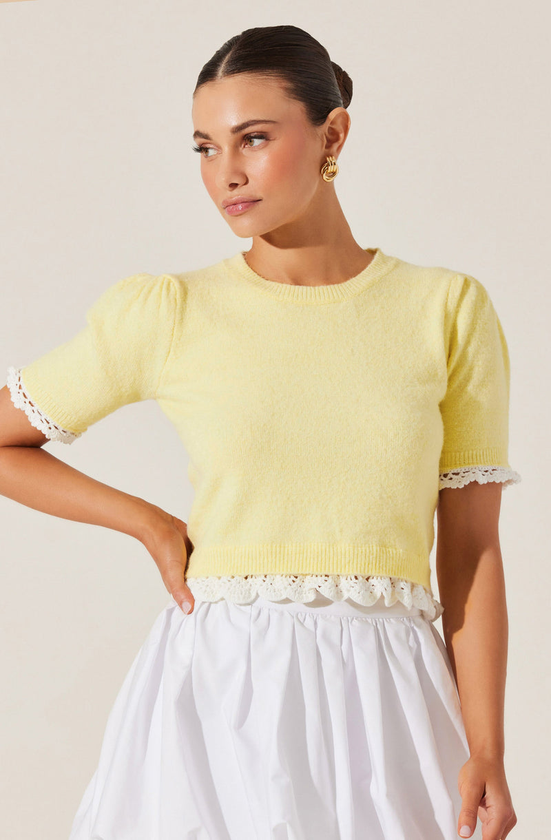 A woman is wearing a light yellow, short-sleeved knit top with puffed sleeves that have lace trim at the cuffs. The top has a scalloped lace hem at the bottom. She is also wearing a white, high-waisted, gathered skirt. She has her hair styled in a sleek bun and is accessorized with large, gold hoop earrings.