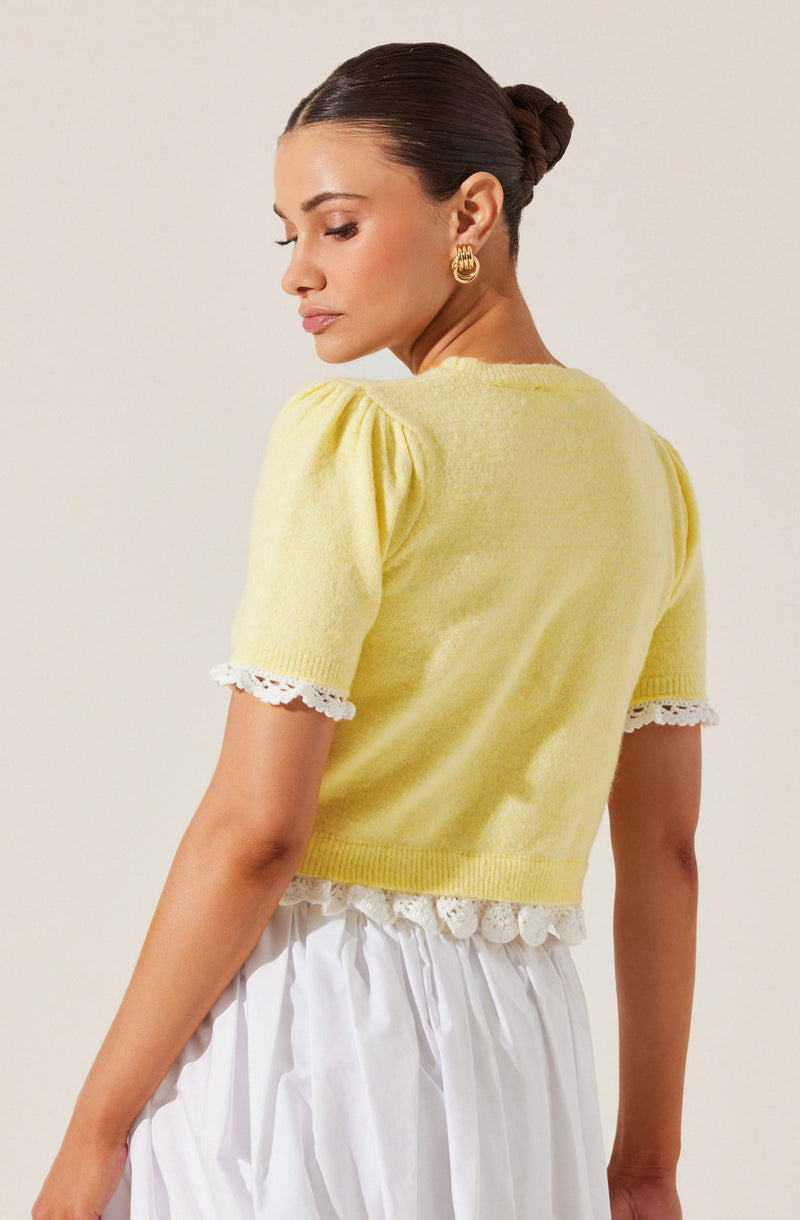 A woman is wearing a light yellow short-sleeved sweater with puffed sleeves that have white lace trim at the cuffs and a scalloped lace trim at the hem. She pairs it with a white skirt that features a gathered or pleated design. She has her hair styled in a sleek bun and is accessorized with gold hoop earrings.