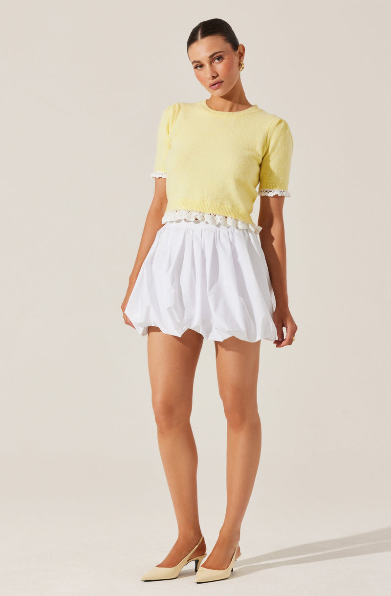 A woman is wearing a short-sleeved, light yellow knit top with lace trim on the sleeves and hem. She pairs it with a white, high-waisted, balloon-style skirt that has a gathered, puffed appearance. She completes her outfit with beige pointed-toe high heels.