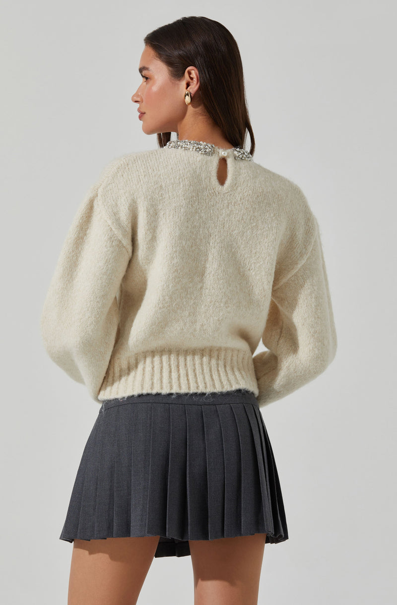 The person is wearing a cream-colored, fuzzy knit sweater with a small keyhole opening at the back, fastened with a decorative button or clasp at the neckline. The sweater has a ribbed hem and cuffs. They are also wearing a high-waisted, dark gray pleated skirt.