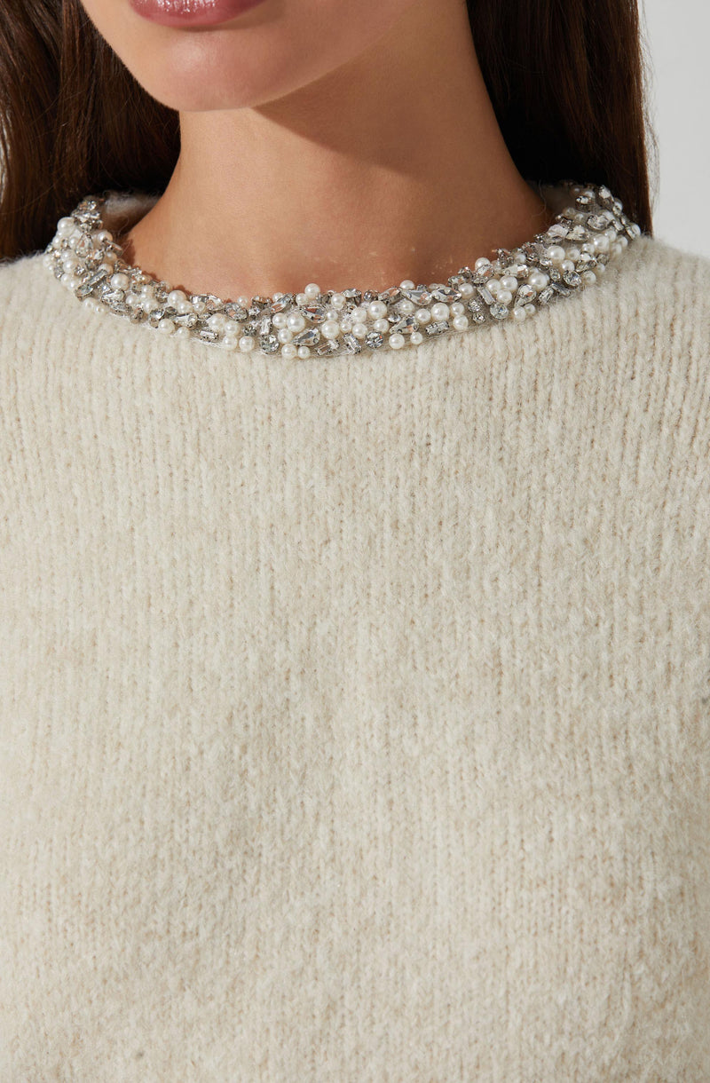 A close-up of a woman wearing a cream-colored, textured knit sweater with a round neckline embellished with a dense cluster of white pearls and sparkling rhinestones.