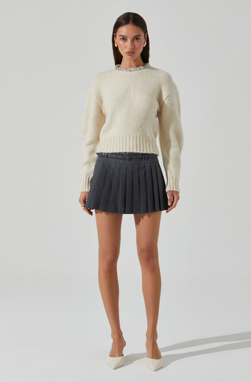 A woman is wearing a chunky, cream-colored knit sweater with long sleeves and a ribbed hem. She has paired it with a dark gray pleated mini skirt with a waistband, and is wearing white high-heeled shoes. She accessorizes with large gold earrings and has long, straight dark hair.