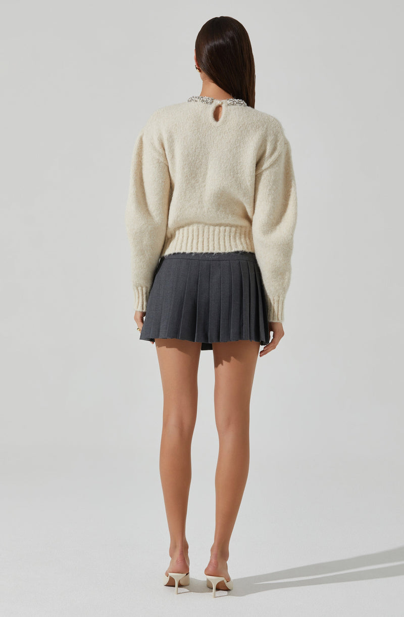A woman is wearing a cream-colored, fuzzy knit sweater with long sleeves and a small keyhole opening at the back near the neckline. She pairs the sweater with a high-waisted, dark gray pleated mini skirt. She is also wearing light-colored high-heeled shoes.