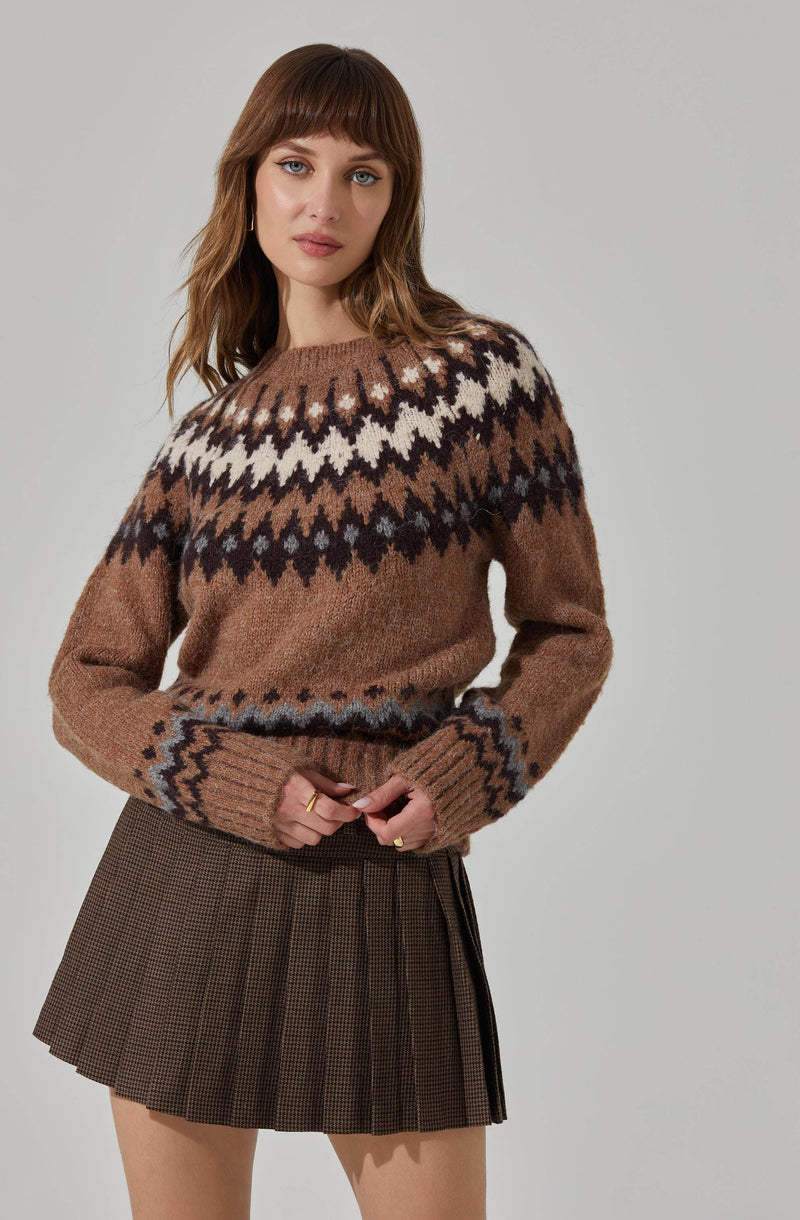A woman is wearing a cozy, oversized sweater with a patterned design featuring white, black, and shades of brown across the chest and cuffs. The sweater is brown with a fair isle pattern around the yoke and sleeve cuffs. She pairs it with a dark brown plaid mini skirt with pleats.