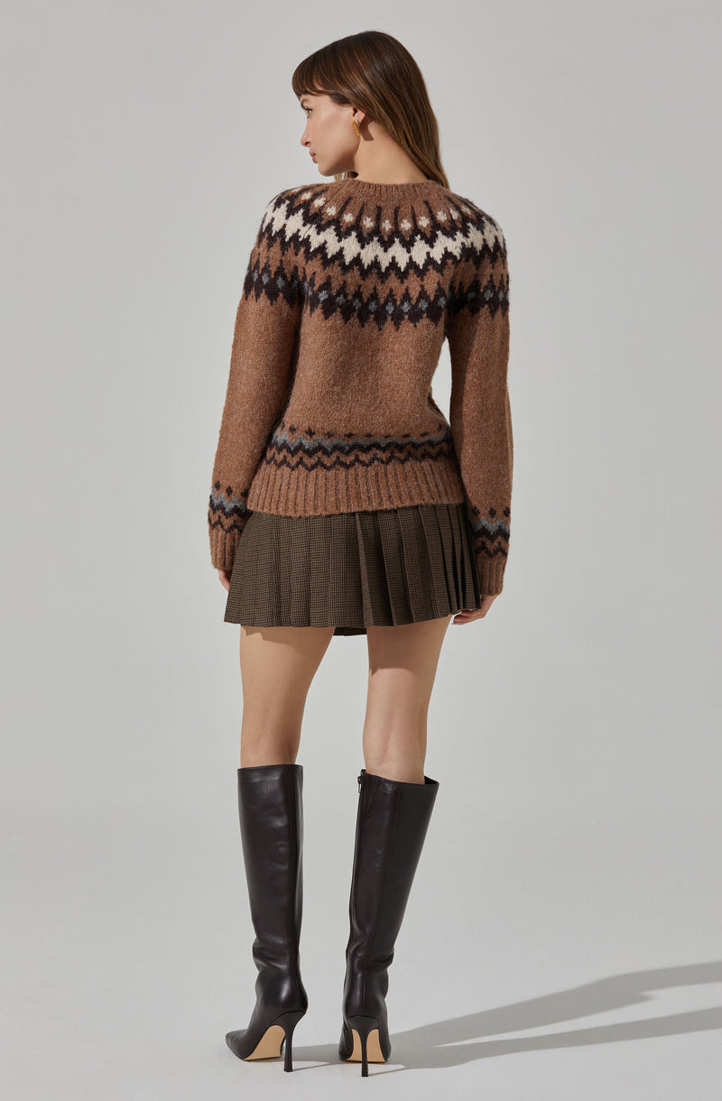 A woman is wearing a multicolored knit sweater with a pattern featuring zigzags and geometric shapes in shades of brown, black, white, and beige. She pairs it with a short, pleated skirt in a brown plaid pattern and black knee-high leather boots with high, stiletto heels.