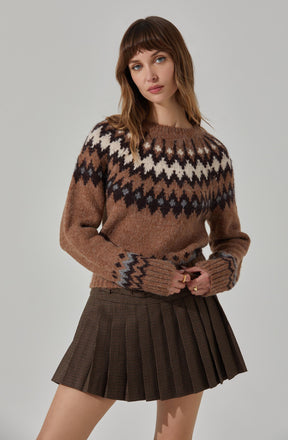 A young woman is wearing a cozy, knit sweater with a geometric pattern in shades of brown, black, white, and gray around the yoke and cuffs. She pairs it with a high-waisted, pleated mini skirt in a brown plaid pattern.