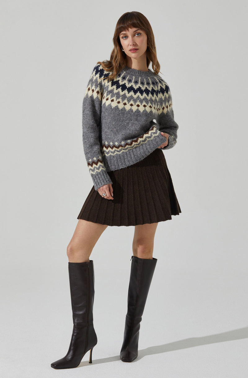 A woman is wearing a knit sweater featuring a multicolored pattern with shades of gray, cream, navy, and brown, along with a dark brown pleated skirt and knee-high black leather boots with high heels.