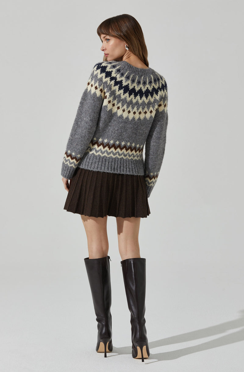 A woman is wearing a cozy, oversized knit sweater with a Fair Isle pattern in shades of gray, navy, cream, and brown. She pairs it with a black, pleated, short skirt and tall, black leather high-heeled boots that reach her knees.