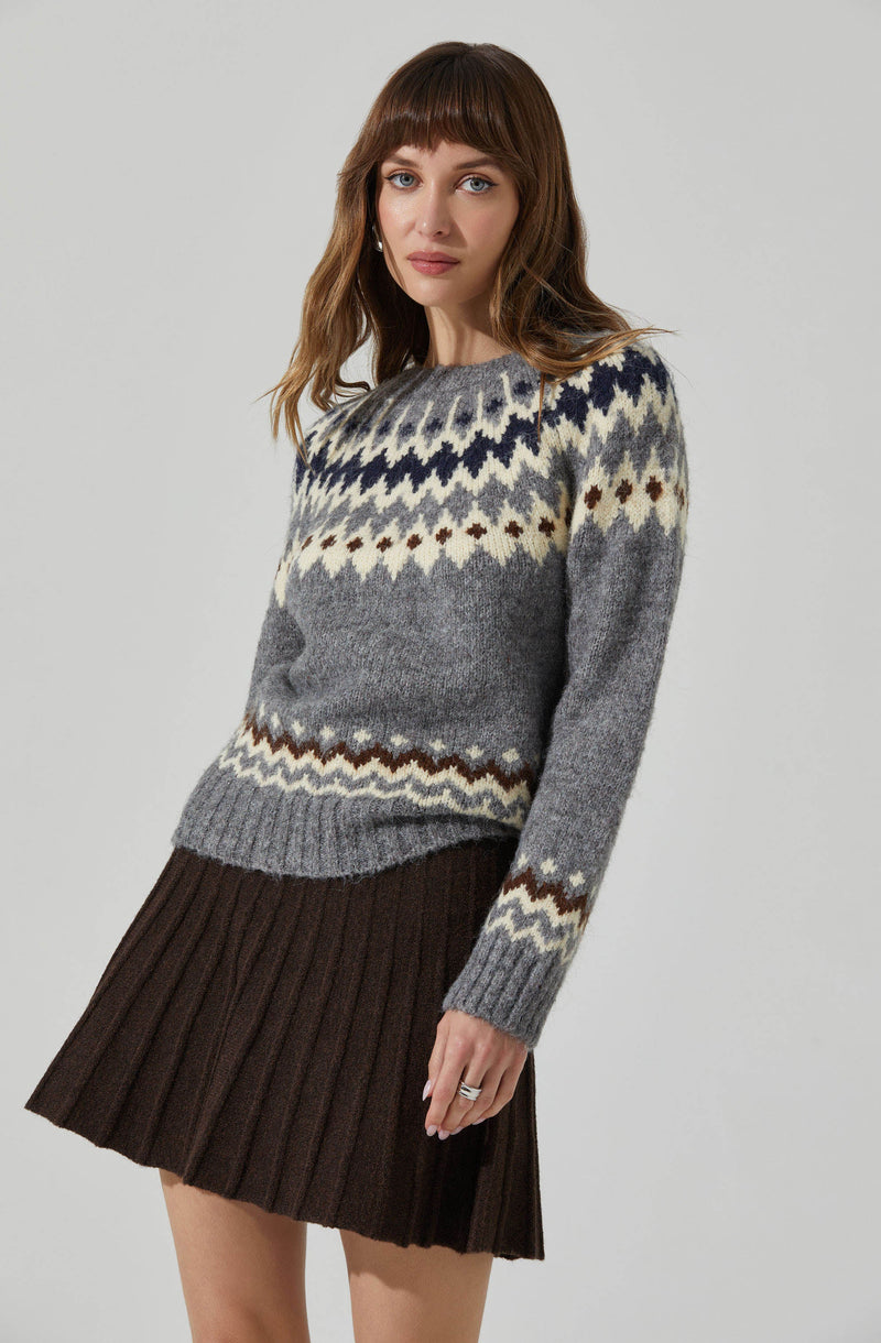 A woman is wearing a cozy knit sweater with a traditional Fair Isle pattern in shades of cream, navy blue, gray, and brown. The sweater has a crew neckline and long sleeves. She pairs it with a dark brown, ribbed, pleated skirt.