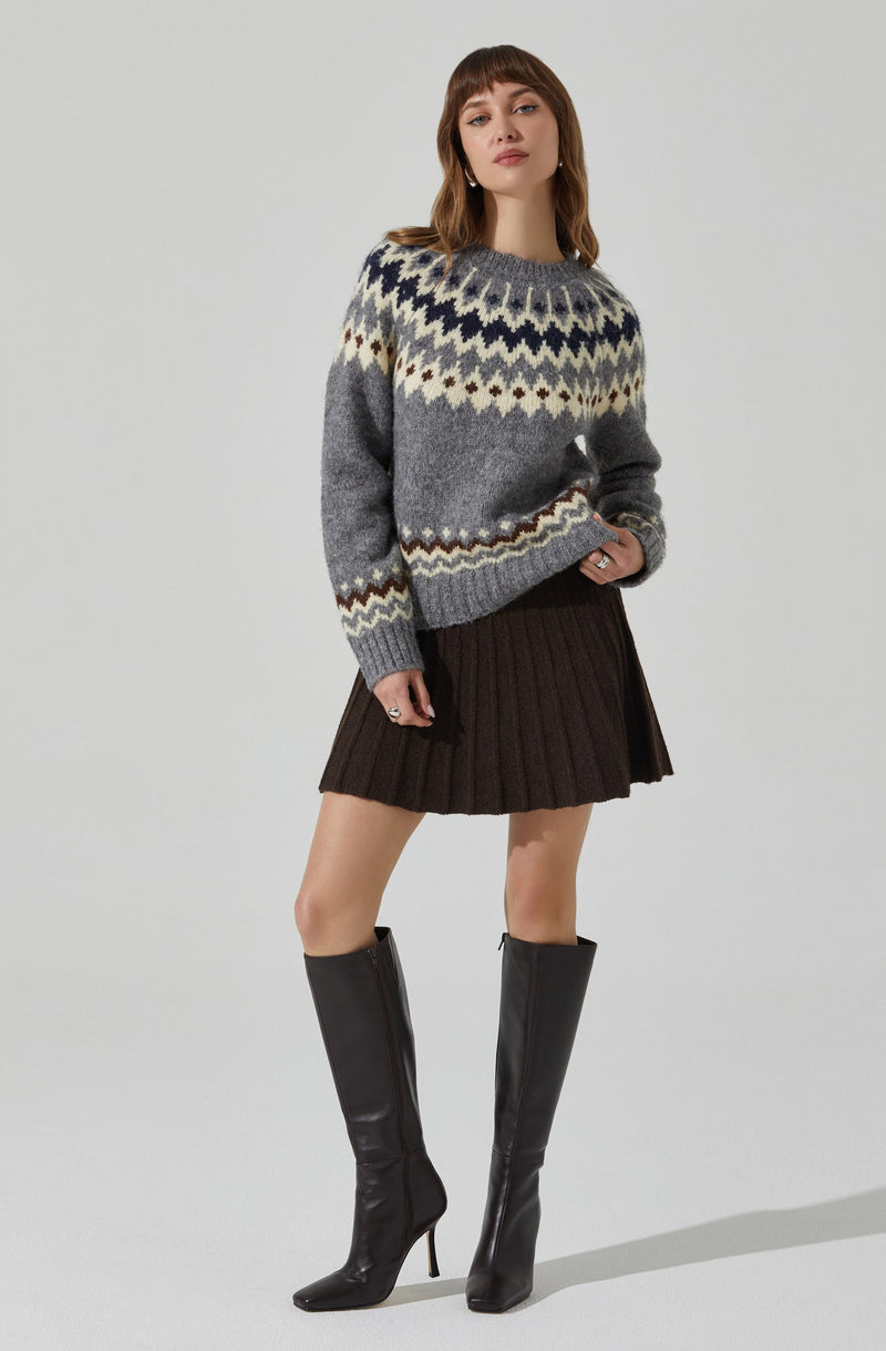 A woman is wearing a Nordic-patterned sweater in shades of gray, cream, and navy blue, with a ribbed collar, cuffs, and hem. She is also dressed in a dark brown, pleated, short skirt and black knee-high heeled boots.