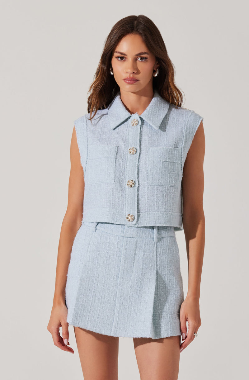 A young woman is wearing a light blue sleeveless cropped top with a collar and four decorative buttons down the front, paired with a matching high-waisted, A-line mini skirt. The outfit is made of a textured, woven fabric.