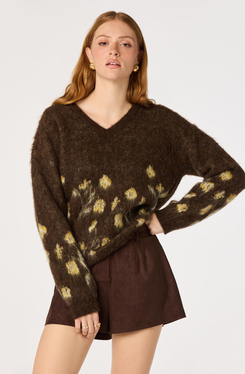 Woman wearing a brown fuzzy sweater with yellow floral patterns and dark brown shorts, with one hand on her hip.