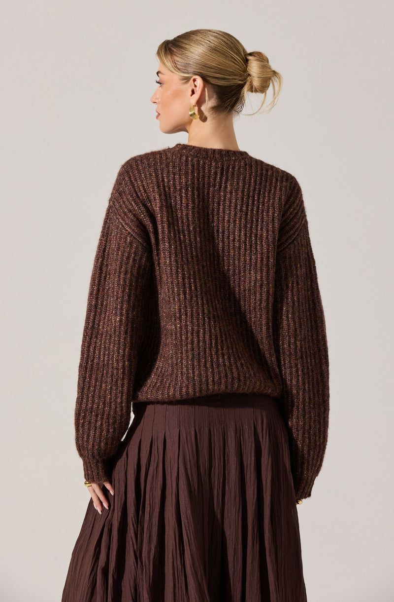 A woman is wearing a loose-fitting, long-sleeved sweater with a multicolored ribbed pattern in shades of brown, maroon, and hints of other earthy tones. The sweater has a round neckline and extends slightly below her waist. She pairs it with a high-waisted, pleated skirt in a matching dark brown color.