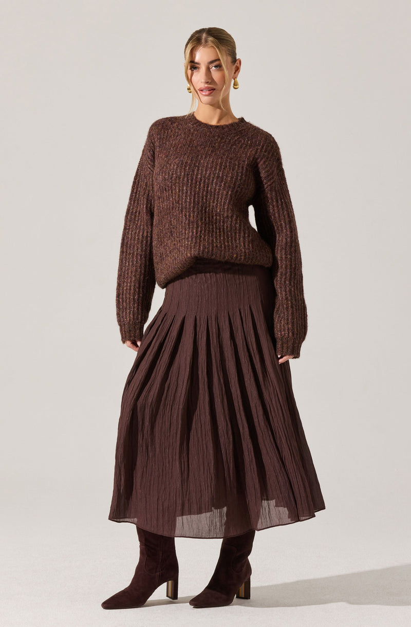A woman is wearing a dark brown, chunky knit sweater with a textured pattern. She pairs it with a long, pleated skirt in a matching brown shade and dark brown, heeled ankle boots made of suede. She accessorizes with gold hoop earrings.