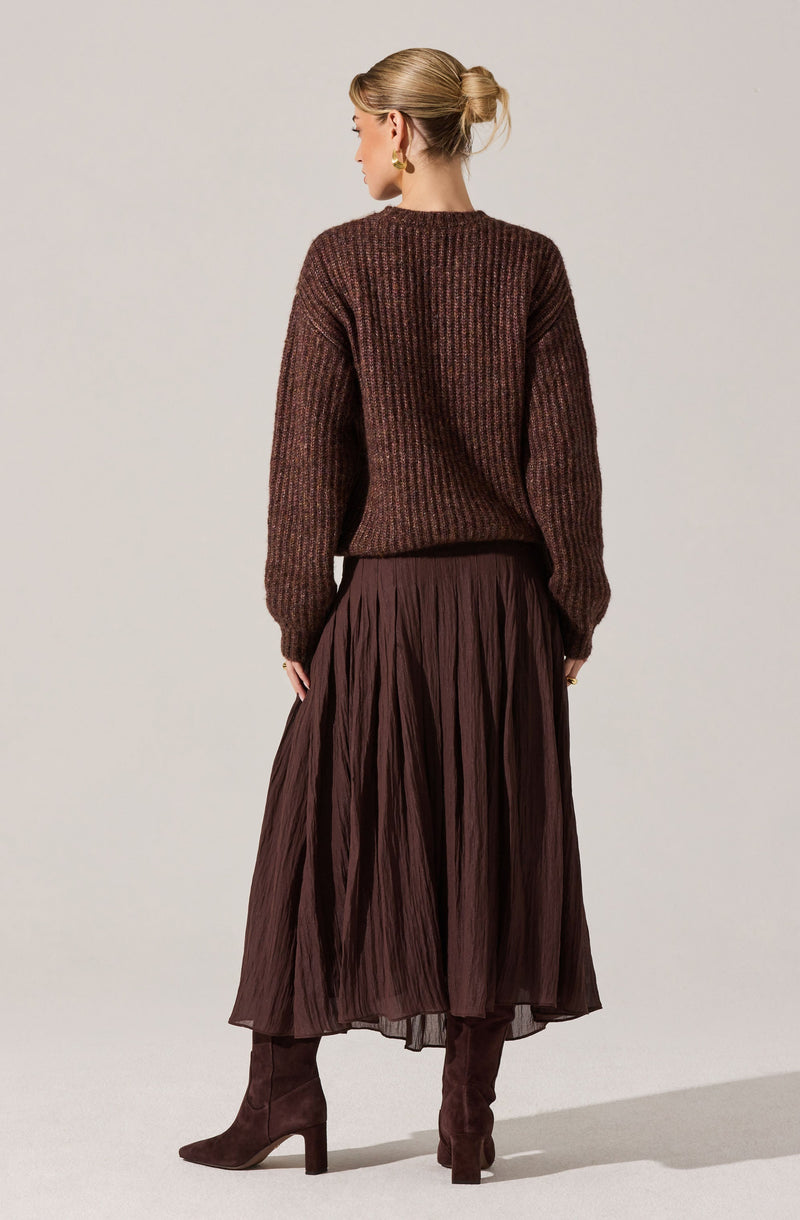 A woman is wearing a brown, chunky knit sweater with a ribbed texture, paired with a long, pleated, dark brown skirt. She has on brown suede high-heeled boots and gold earrings, with her blonde hair styled in a low bun.