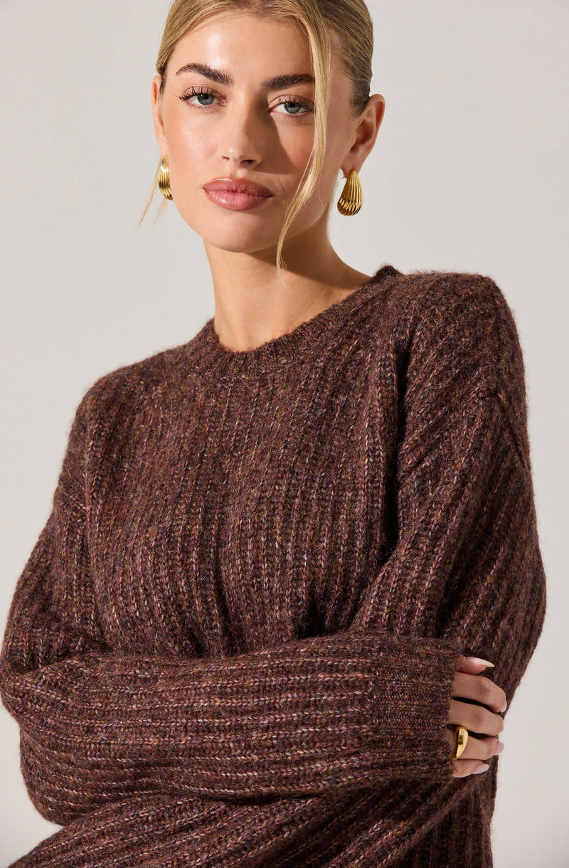 A woman is wearing a cozy, oversized sweater with a marled brown, purple, and pink color palette. The sweater has a ribbed, textured knit pattern and long sleeves. She is accessorized with large, gold, Egyptian-style hoop earrings and a gold ring on her right hand. Her hair is styled in a sleek, blonde updo, and her makeup is natural with a focus on her eyebrows and lips.