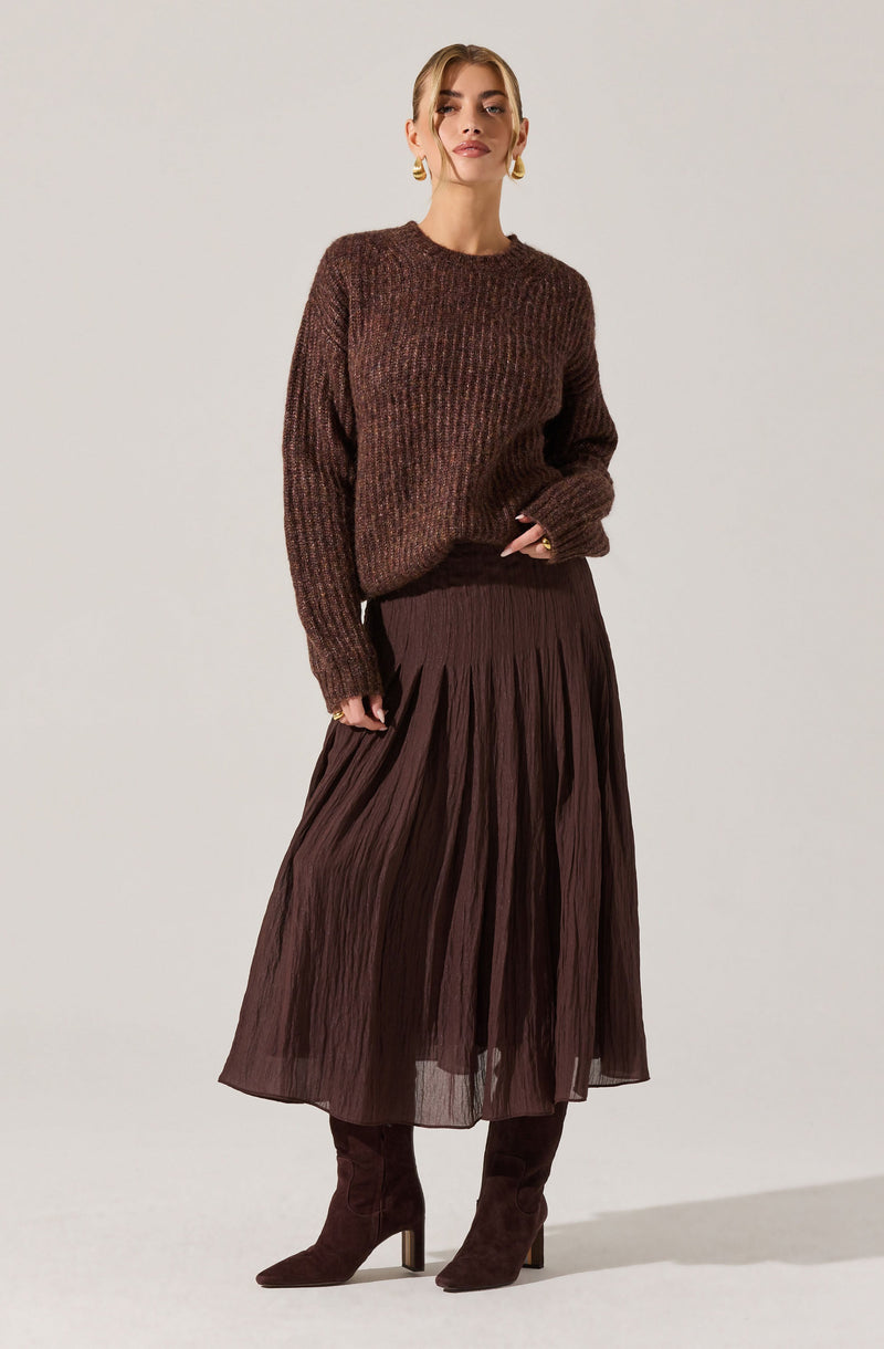 A woman is wearing a loose-fitting, textured, brown knitted sweater with a round neckline and long sleeves. She pairs it with a dark brown, pleated, midi-length skirt made of a lightweight, semi-sheer fabric. She completes the outfit with matching dark brown suede high-heeled boots and gold hoop earrings.