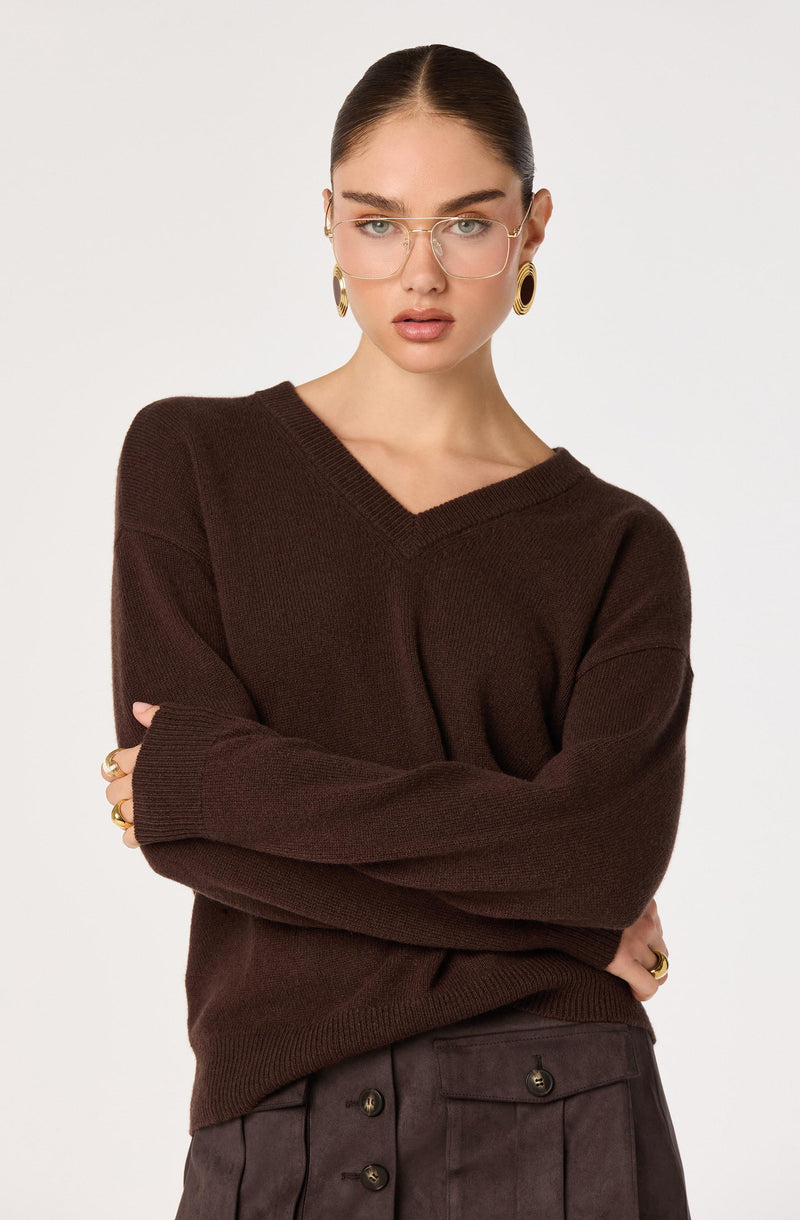 Woman in a dark brown V-neck sweater with rolled-up sleeves, wearing glasses and gold jewelry.