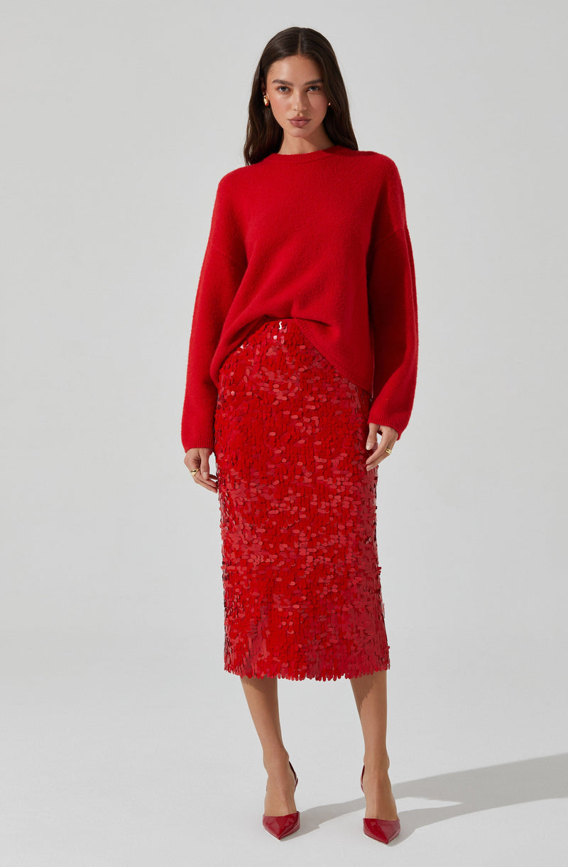 A woman is wearing a bright red, oversized, long-sleeve knit sweater paired with a matching red, high-waisted, midi-length skirt that features a textured, sequined pattern. She completes the outfit with red pointed-toe high heels.