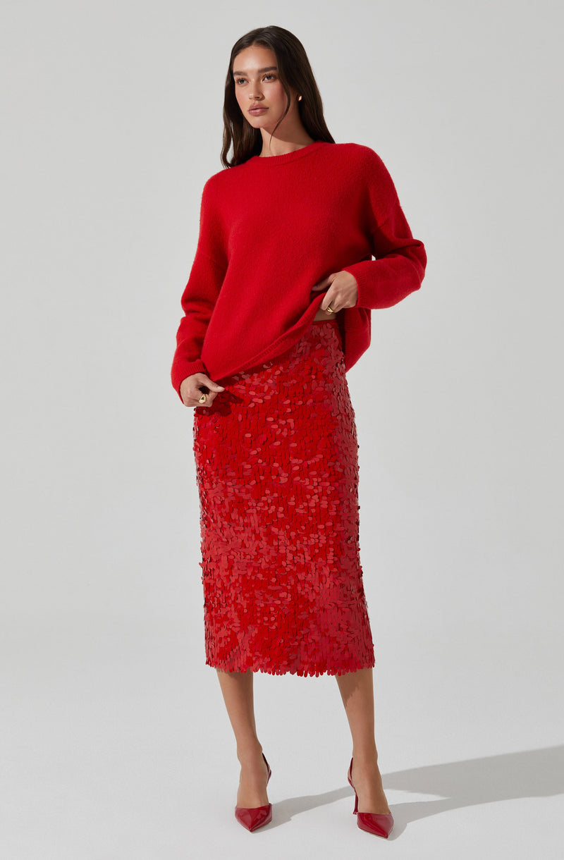 A woman is wearing a bright red, long-sleeved, oversized sweater paired with a red textured midi skirt that has a floral or abstract pattern. She completes her outfit with red pointed-toe heels.