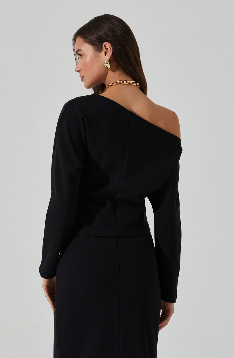 A woman is wearing a black, off-the-shoulder top with long sleeves, paired with a matching black skirt. The top drapes elegantly, exposing her left shoulder. She accessorizes with a chunky gold chain necklace and gold hoop earrings.