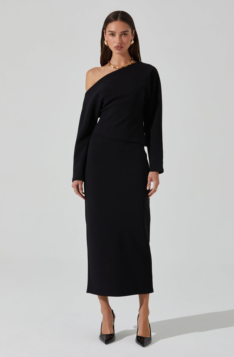 A woman is wearing an elegant, all-black outfit consisting of a long skirt and a matching off-the-shoulder top. The top features a relaxed fit with one shoulder exposed, while the skirt is high-waisted and flows down to her ankles. She accessorizes with gold hoop earrings, layered necklaces, and rings, completing her sophisticated look with black pointed-toe high heels.