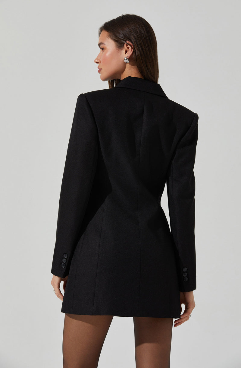 A woman is wearing a tailored, black blazer-style dress with long sleeves that have three buttons on each cuff. The dress features a structured fit with defined seams, a notched collar, and a hemline that falls above the knees.