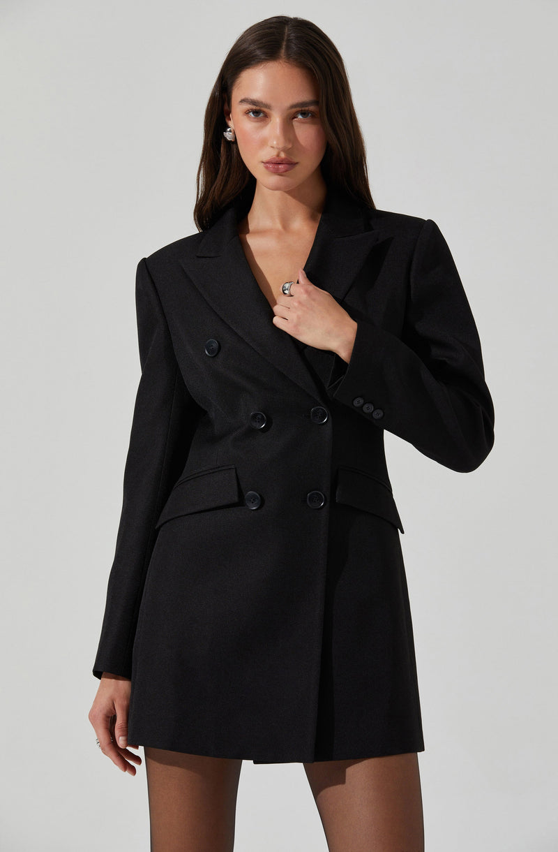 A woman is wearing a black, double-breasted blazer with a tailored fit. The blazer features peak lapels, long sleeves with four buttons on each cuff, and two flap pockets on the front. She is also wearing sheer black tights and has earrings and a ring as accessories.