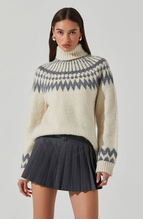 A woman is wearing a cream-colored, chunky knit turtleneck sweater with a gray geometric pattern around the yoke, cuffs, and hem. She pairs it with a high-waisted, black pleated mini skirt featuring thin vertical stripes.