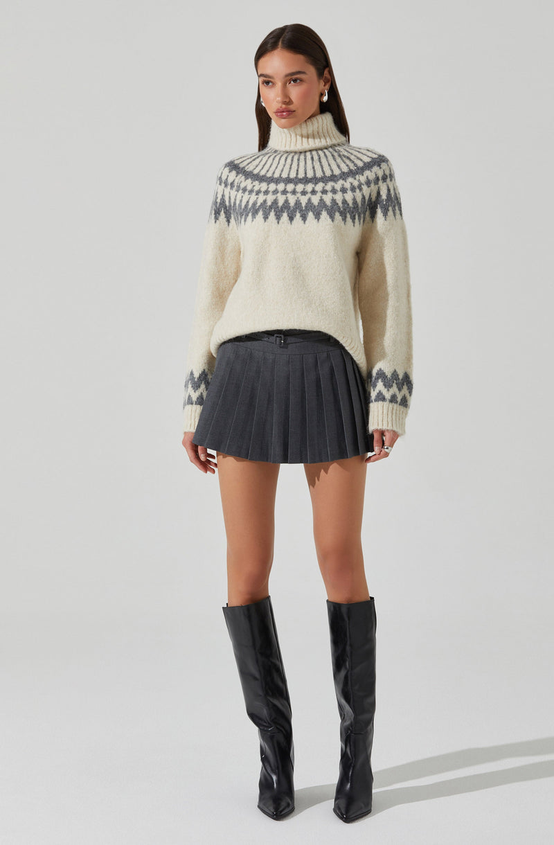 A woman is wearing an oversized, cream-colored turtleneck sweater with a gray and white geometric pattern around the yoke and sleeves. She pairs it with a high-waisted, dark gray pleated mini skirt. She completes the outfit with black, knee-high boots with a shiny finish.