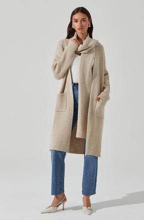 A woman is wearing a long, beige, ribbed knit cardigan with large front pockets and a matching knit scarf draped over her shoulders. She has on a white top underneath and blue high-waisted jeans. Her footwear consists of white pointed-toe heels with ankle straps.