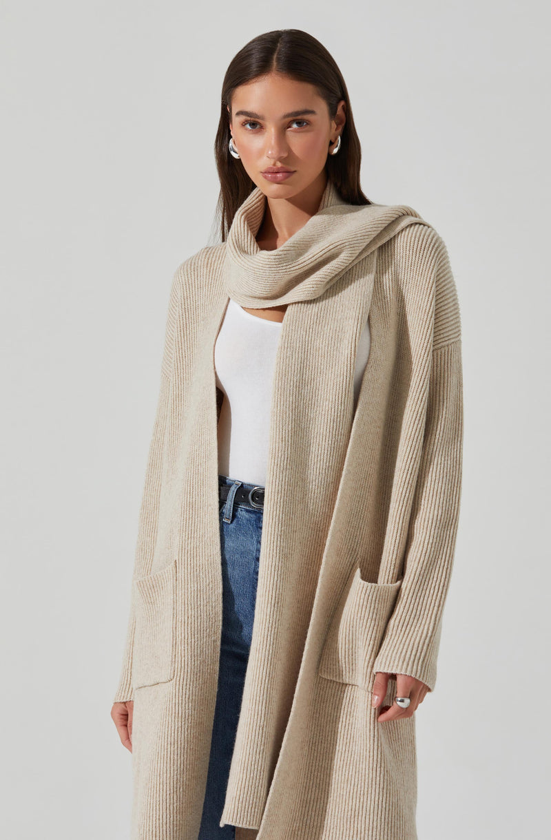 A woman is wearing a long, beige, ribbed knit cardigan with large front pockets. She has a matching beige scarf draped around her shoulders, a white top underneath, and blue jeans.