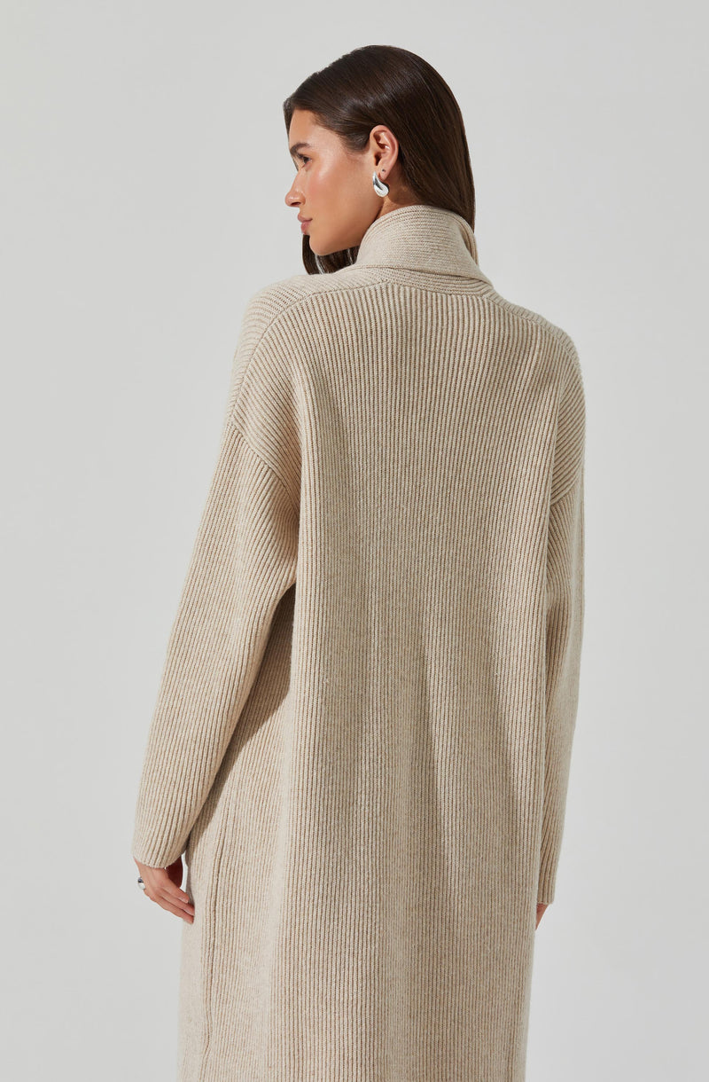A woman is wearing a long, beige, ribbed knit coat with a high collar. The coat appears cozy and oversized, with relaxed fit and dropped shoulders. She has dark, straight hair and is accessorized with silver hoop earrings.