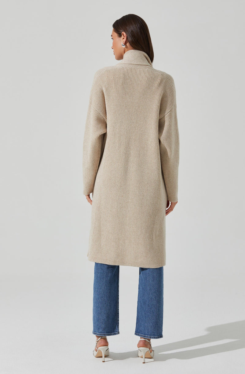 A woman is wearing a long, beige, ribbed knit cardigan with a high collar, paired with straight-leg blue jeans and beige heeled sandals.