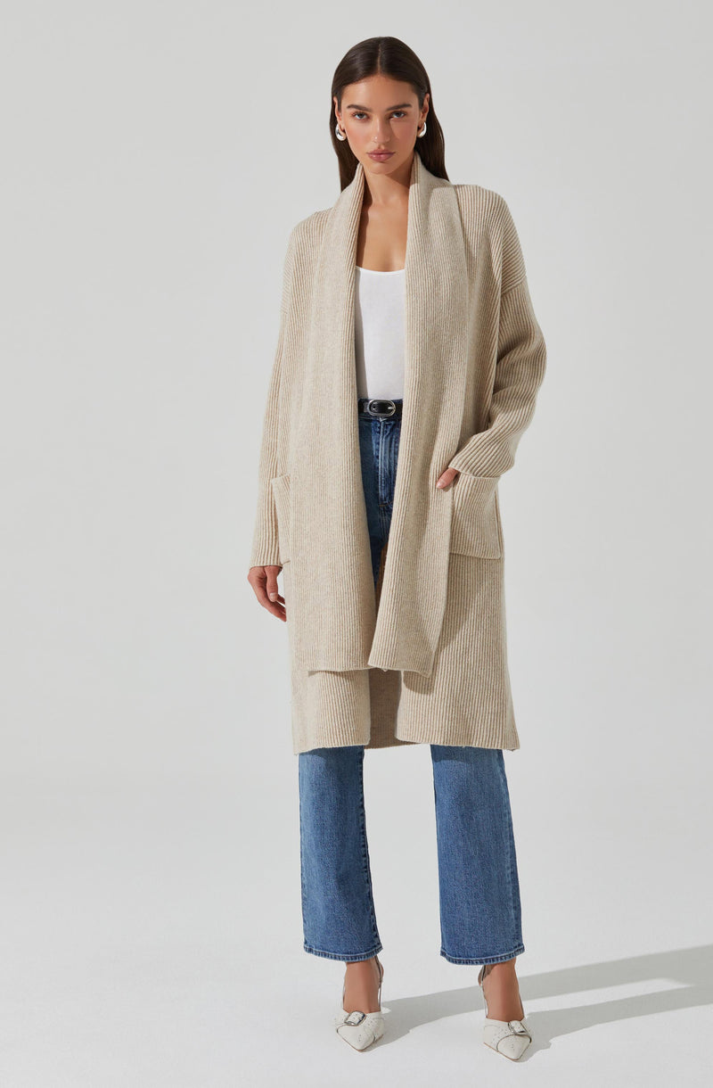 A woman wearing a long, beige, ribbed knit cardigan with large front pockets, paired with a white top, high-waisted blue jeans, and white pointed-toe shoes.
