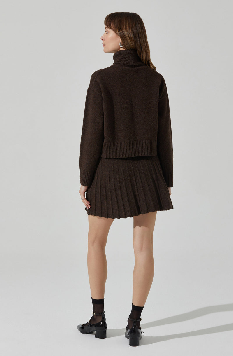 A woman is wearing a dark brown, chunky knit turtleneck sweater with a loose fit and ribbed hem. She pairs it with a matching dark brown pleated mini skirt. She is also wearing black ankle socks and black heeled shoes with ankle straps.